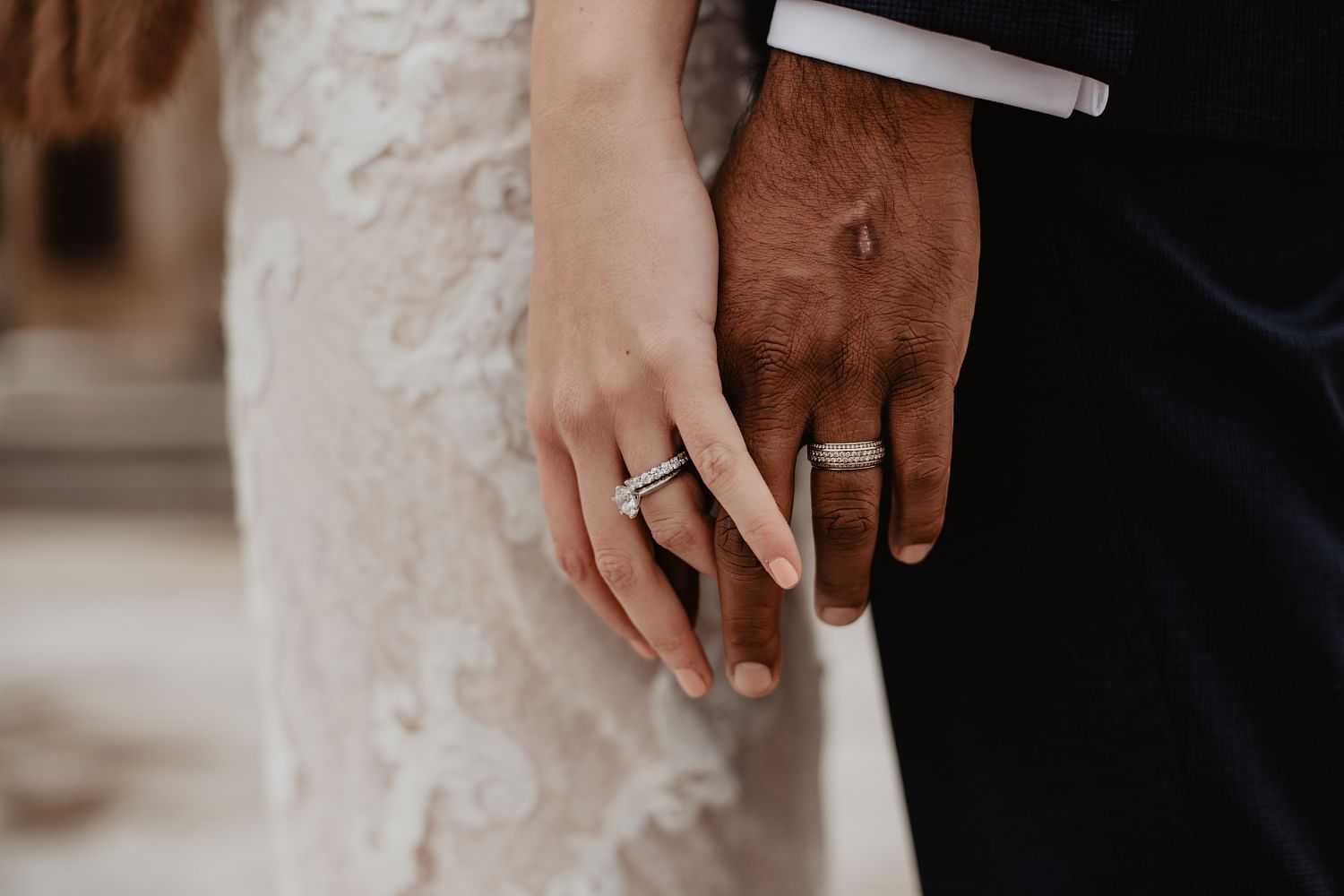 Close-up of a couple's hands with wedding rings, one wearing a lace dress, the other a suit.